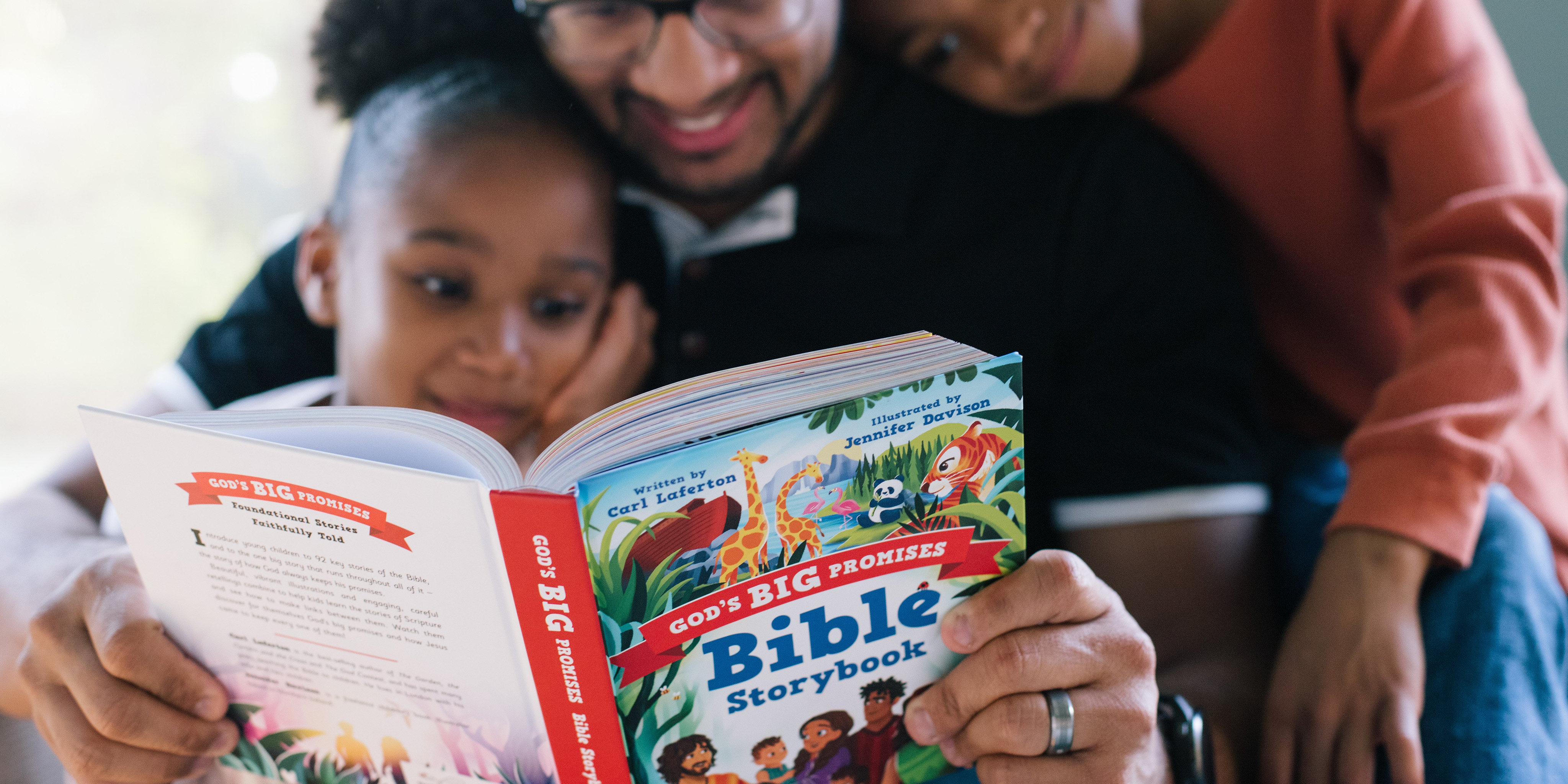 Photograph of two sons and a father. The father is seated and reading God's Big Promises Bible Storybook. The sons are standing on either side of the father looking at the Bible Storybook.
