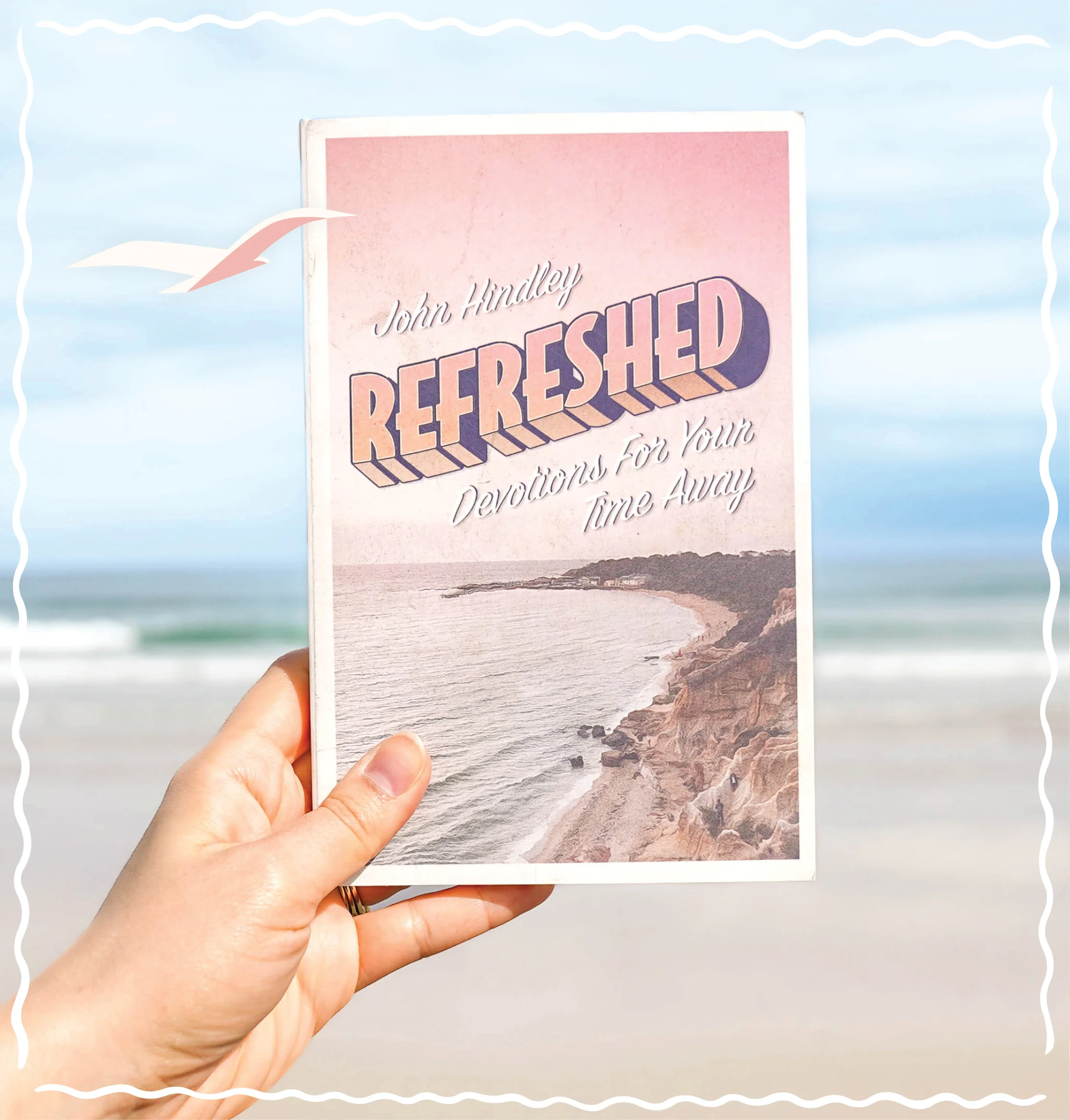 Photograph featuring a person holding up Refreshed: Devotions for Your Time Away with a beach scene in the background