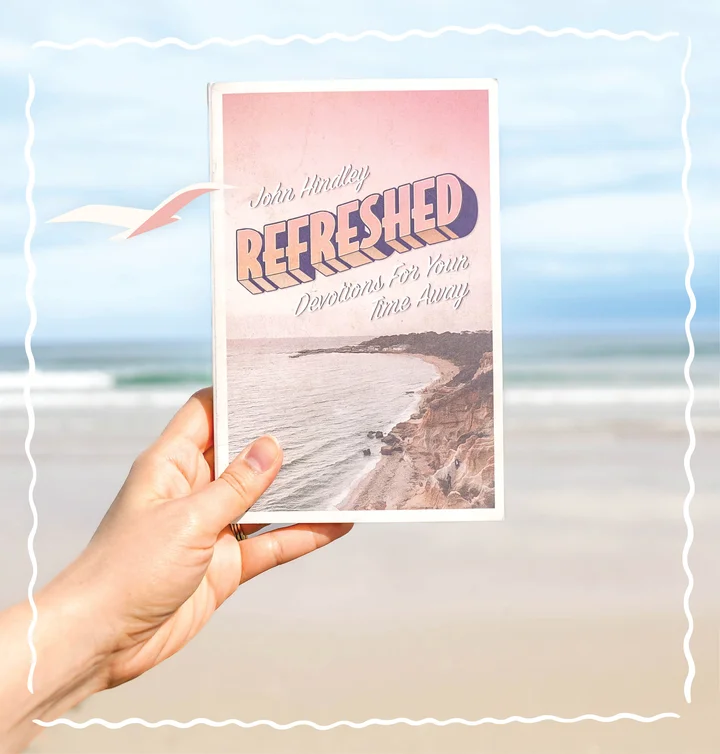 Photograph featuring a person holding up Refreshed: Devotions for Your Time Away with a beach scene in the background